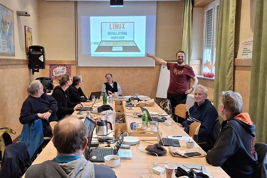 Linux-Installations-Workshop am 20.12.2025 im Clubraum des ADFC Münsterland. Auf dem großen Gruppentisch im Clubraum des ADFC Münsterland stehen viele geöffnete Laptops. An dem Tisch sitzen Menschen. Auf der Leinwand ist eine Zeichnung von einem Laptop zusehen auf dem Bildschirm steht : Linux, Linux-Stammtisch Münster, Installations-Workshop, ADFC-Münsterland. Auf einem USB-Stick steht Linux.
